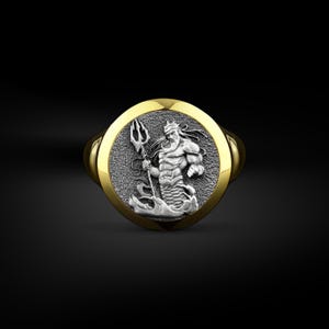 Pode incluir: Anel dourado com um relevo prateado detalhado de uma figura musculosa segurando um tridente. A figura está dentro de uma moldura circular texturizada. O anel tem uma banda polida e arredondada.