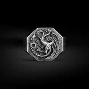 Pode incluir: Anel prateado com uma face octogonal com um design detalhado de dragão. O dragão é retratado com asas e uma cauda curva, rodeado por detalhes ornamentados. O anel tem um acabamento em metal escovado.