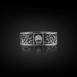 May include: A silver ring with a lion's head in a rectangular frame. The ring features ornate floral scrollwork around the band. The ring is displayed on a reflective black surface.
