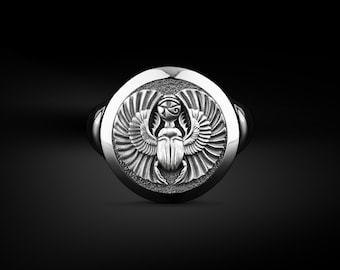 May include: A silver ring with an engraved scarab beetle design, complete with wings and an Eye of Horus. The ring has a polished finish and is set against a dark background, accentuating the intricate details.