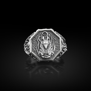 Puede incluir: Anillo plateado con una cara octogonal que presenta un relieve detallado de Anubis, el dios egipcio del más allá, con llamas alrededor de la cabeza. El anillo tiene una banda ornamentada y texturizada con diseños florales.