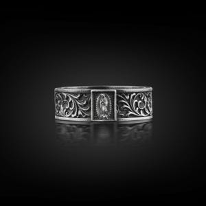 Könnte beinhalten: Ein silberner Ring mit einem aufwendigen floralen Muster und einem zentralen rechteckigen Feld mit einer religiösen Figur. Das Band des Rings ist mit wirbelnden Mustern versehen, was einen Vintage-Look erzeugt. Der Ring wird vor einem dunklen Hintergrund präsentiert.