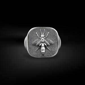 Op de afbeelding: Een zilveren ring met een vierkante kop, met een gedetailleerd reliëf van een bij. De bij heeft uitgespreide vleugels en een gesegmenteerd lichaam. De ring staat tegen een zwarte achtergrond.
