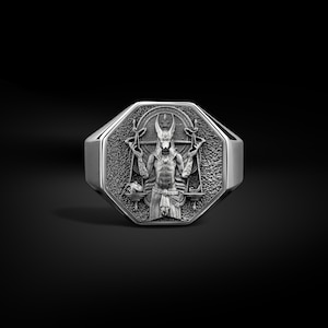 May include: A silver ring with an octagonal face. The face features a detailed relief of a figure holding scales, with an Egyptian theme. The ring has a polished finish and a thick band, suitable for a man or woman.