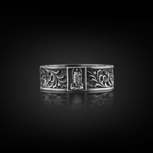 May include: A silver ring with an ornate floral design and a central rectangular panel featuring a figure. The ring has a wide band with detailed scrollwork and a dark, reflective background.