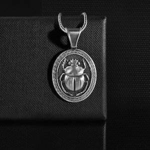 May include: Silver-toned pendant featuring a detailed scarab beetle design within an oval frame. The pendant hangs from a matching chain, showcasing an ancient Egyptian theme. The beetle is the focal point, surrounded by a decorative border.