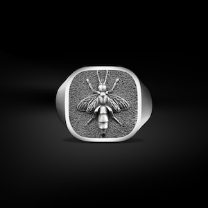 Op de afbeelding: Een zilverkleurige ring met een gedetailleerd bijenontwerp. De bij is gecentreerd op een vierkante face met een getextureerde achtergrond. De ring heeft een gepolijste, afgeronde band. De vleugels van de bij zijn gespreid en het lichaam is gesegmenteerd.