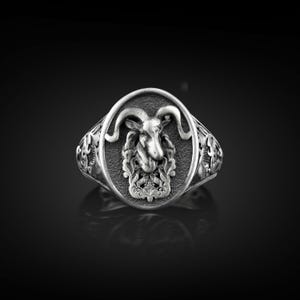 May include: A silver-colored ring featuring a detailed ram's head within an oval frame. The ring has ornate scrollwork on the band and around the oval. The ram's head has curved horns and a detailed face. The ring is likely made of metal.