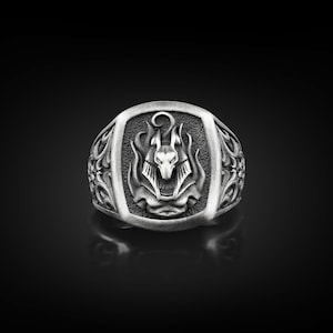 Puede incluir: Un anillo de plata con un diseño de Anubis. El anillo muestra al dios egipcio Anubis, con llamas y patrones detallados. El anillo tiene una cara rectangular y una banda detallada.