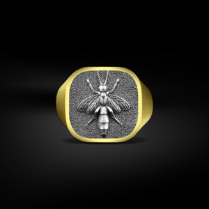 Op de afbeelding: Een goudkleurige ring met een vierkante voorkant met een gedetailleerd zilverkleurig bijenontwerp. De bij heeft uitgespreide vleugels en ingewikkelde lichaamsdetails. De ring heeft een gepolijste afwerking.