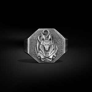 Puede incluir: Anillo plateado con una cara octogonal. El anillo presenta un relieve detallado de una figura con cabeza de chacal, posiblemente Anubis, rodeada de llamas. El anillo tiene una superficie texturizada y un fondo oscuro.