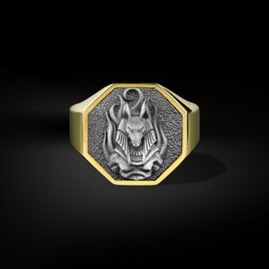 Puede incluir: Anillo dorado con una cara octogonal que presenta un detallado diseño de Anubis plateado. La figura de Anubis está rodeada de llamas, sobre un fondo texturizado. El anillo tiene un acabado pulido.