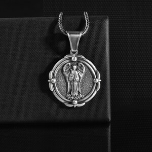 May include: A silver pendant necklace with an angel design. The circular pendant has an elaborate border and a textured backdrop. The angel is shown with wings and a flowing robe. The necklace chain is a textured, woven style.