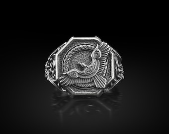 May include: A silver-toned ring with an octagonal face featuring a detailed bird design. The bird is depicted with outstretched wings within a circular frame, surrounded by ornate patterns. The ring has a textured finish and is set against a dark background.