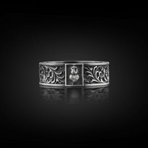 May include: A silver ring with an elaborate floral pattern. The ring has a central rectangular panel with a heart and cross emblem. The band is approximately 1.3 cm wide, with detailed scrollwork. The ring is set against a dark background.