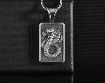 May include: A silver pendant featuring a detailed cobra design within a rectangular frame. The snake is coiled, with its hood raised and fangs bared. The pendant hangs from a silver chain, set against a dark background.