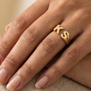 Peut inclure: Une bague dorée avec les lettres "KS" en gras. La bague est portée au doigt, et les lettres sont bien visibles. La bague semble être en métal et a une finition polie.