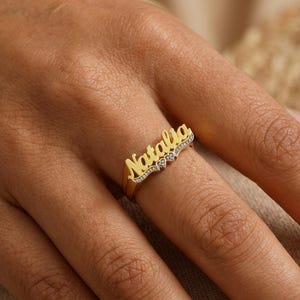 May include: A gold-colored ring with the name "Natalia" in cursive script. The ring features small, clear gemstones and a heart-shaped design. The ring is worn on a finger.