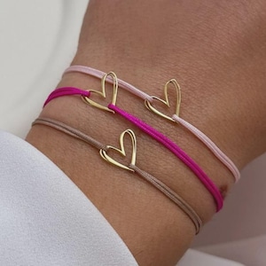 Könnte beinhalten: Drei zarte Armbänder mit goldenen Herzanhängern. Die Armbänder haben dünne, farbige Kordeln in Rosa, Magenta und Taupe. Die goldenen Herzanhänger sind auf jedem Armband zentriert und schaffen ein minimalistisches und elegantes Design.