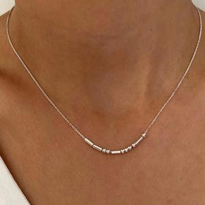 Op de afbeelding: Een delicate zilveren ketting met een reeks kleine, cilindrische en bolvormige kralen. De ketting wordt om de nek gedragen en rust op het sleutelbeen. De achtergrond is een witte blouse.