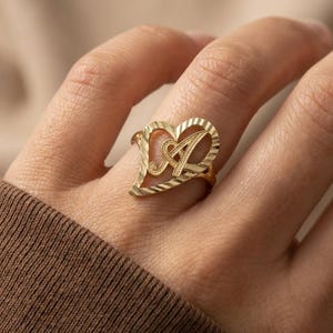May include: A gold ring with a heart-shaped design and the letter "A" in the centre. The ring has a textured surface and is worn on a finger. The ring is a piece of jewellery.