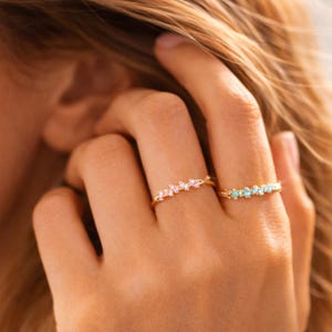 Puede incluir: Dos anillos dorados con detalles de piedras preciosas. Uno tiene piedras rosas, el otro azules. Los anillos se muestran en una mano. Las piedras están engastadas en oro.