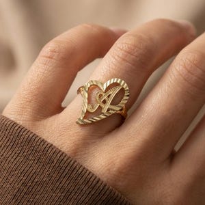 May include: A gold-colored ring featuring a heart-shaped design with the letter "A" in the center. The ring has a textured surface and is worn on a finger. The ring is a piece of jewelry.