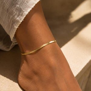 May include: A thin, gold-toned herringbone chain anklet resting around the ankle. The jewelry features a flat, flexible link design that catches the light, creating a smooth, reflective surface that sits flush against the skin in a simple, continuous loop.