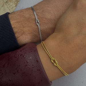 May include: A close-up of two wrists, each adorned with a delicate knot bracelet. One bracelet is silver, the other gold. The bracelets are the focal point, set against a backdrop of clothing in shades of burgundy, navy, and gray.