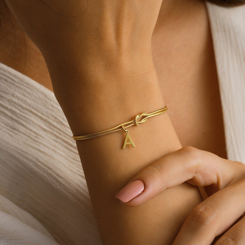 May include: A gold chain bracelet with a knot detail and a letter 'A' charm.