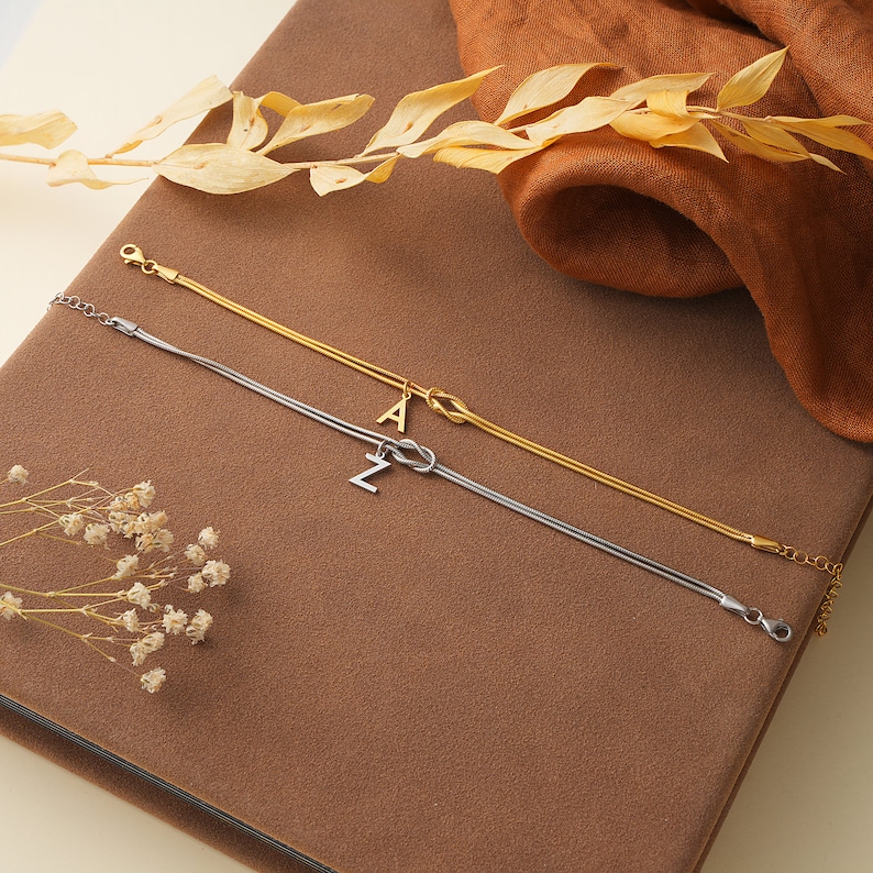 May include: Two delicate necklaces with a knot design. One necklace is gold with a letter 'A' charm, and the other is silver with a letter 'Z' charm.