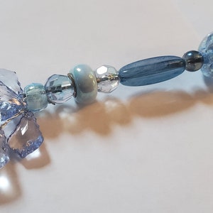May include: A delicate, handmade beaded ornament featuring a cluster of light blue crystal-like shapes and various blue beads. The beads include round, oval, and faceted designs in shades of light and dark blue, strung together with silver wire.