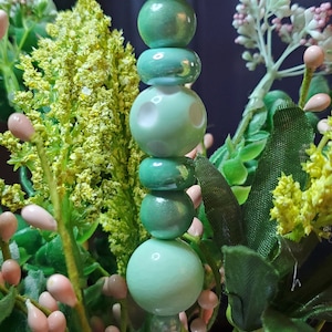 May include: A green glass bead garland with a white bead in the middle, displayed in a glass jar with green and pink artificial flowers.
