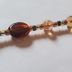 May include: A beaded strand featuring a variety of amber and brown beads. The beads include oval, round, and faceted shapes, with a mix of transparent and opaque finishes. The strand is likely used for jewelry making or as a decorative element.