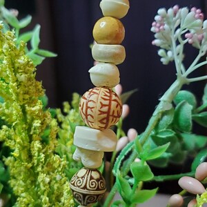 May include: A beaded necklace with brown, white, and orange beads. The beads have intricate patterns and designs. The necklace is hanging from a green plant.