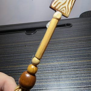 Puede incluir: Un tapón de botella de madera con una parte superior cuadrada con un diseño de rayas de cebra en crema y marrón. El tapón tiene un mango de madera marrón claro con cuentas decorativas de madera en varios tonos de marrón y crema.