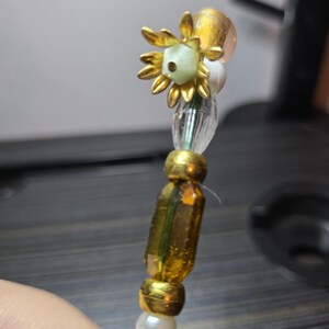 May include: A beaded accessory featuring a gold-colored sunflower charm with a light green center. The beads include gold-colored, clear, and iridescent white beads. The accessory has a vintage aesthetic.