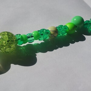 May include: A handmade beaded accessory featuring a variety of green beads. The design includes a large, clear, leaf-shaped bead, a glittery green sphere, and various sizes and shapes of green beads. The accessory is on a white surface.