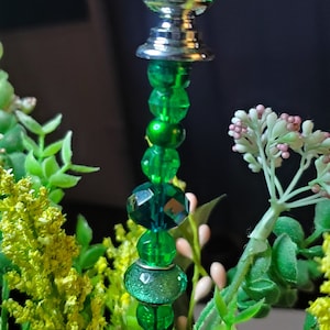 May include: A green glass bottle stopper with a silver base and a beaded stem. The stopper is surrounded by green and yellow artificial foliage.