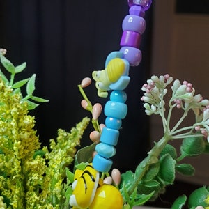 May include: A colorful beaded necklace with a butterfly, bee, and tiger charm. The beads are purple, blue, and yellow. The necklace is surrounded by green leaves and pink flowers.