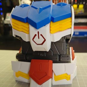 May include: A 3D-printed model of a robot's torso, featuring blue, white, yellow, red, and black components. The design includes a power symbol and geometric shapes, suggesting a futuristic or mechanical theme. The model is assembled with interlocking parts.