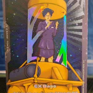 May include: A rectangular trading card featuring a character in a purple suit standing inside a yellow structure. The card displays the text EX BASE TOKEN, EX Base, and At the start of the game, place 1 active EX Base as your shield area's base.
