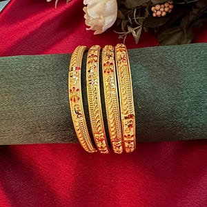 May include: Four gold bangles with detailed floral and geometric designs, featuring red and black enamel accents. The bangles are presented on a green surface against a red backdrop, with a white flower and foliage in the top right corner.