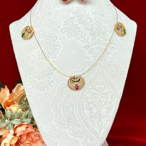 May include: A gold-toned jewelry set featuring a necklace and matching earrings. The necklace has a pendant with a red gemstone, and the earrings also have red gemstones. The set is displayed on a white bust against a red background.