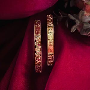 May include: Two gold bangles with intricate floral and scrollwork designs. One bangle features red gemstones and clear accents, while the other showcases a floral motif. The bangles rest on a deep red fabric, suggesting a jewelry display.