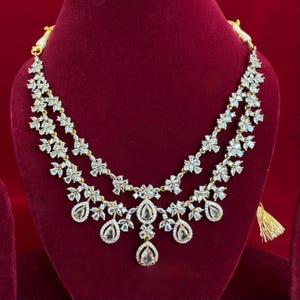 Pu&ograve; includere: Una collana a due fili con un design floreale. La collana presenta pietre chiare e scintillanti incastonate in un metallo color oro. La collana ha un design a pendente a goccia con pietre a forma di goccia. La collana &egrave; esposta su una superficie di velluto rosso.