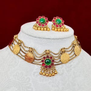 May include: A gold-toned necklace and earring set. The necklace features multiple strands with coin-like pendants and a central floral design with green and red gemstones. The earrings mirror the floral design. The jewelry is displayed on a white bust against a red background.