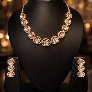 May include: A gold-toned necklace and matching earrings set displayed on a black jewelry bust. The necklace and earrings feature clear, oval-shaped stones surrounded by smaller, sparkling accents. The jewelry is presented against a blurred, warm-toned background.