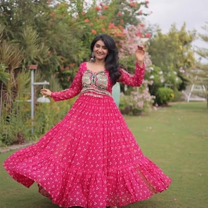May include: A floor-length fuchsia dress with long sleeves and a tiered, flared skirt. The fabric features a small white dot pattern. The bodice displays a decorative panel with colorful floral embroidery and metallic accents at the waistline.