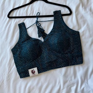 Puede incluir: Un top sin mangas de color verde azulado con cuello en V y detalles de lentejuelas intrincados. El top está colgado de una percha negra sobre un fondo blanco. Una pequeña etiqueta con un diseño floral y la palabra "Anatab" está adherida a la prenda.